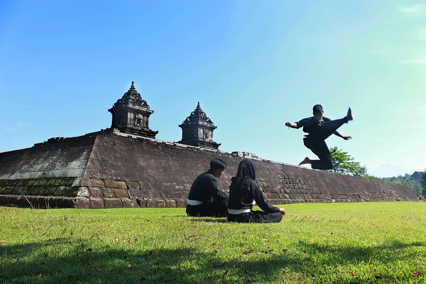 Pratiquant de pencak silat en tenue noire exécutant un coup de pied sauté devant le temple de Candi Barong, près de Yogyakarta, sous le regard de deux élèves assis dans l'herbe