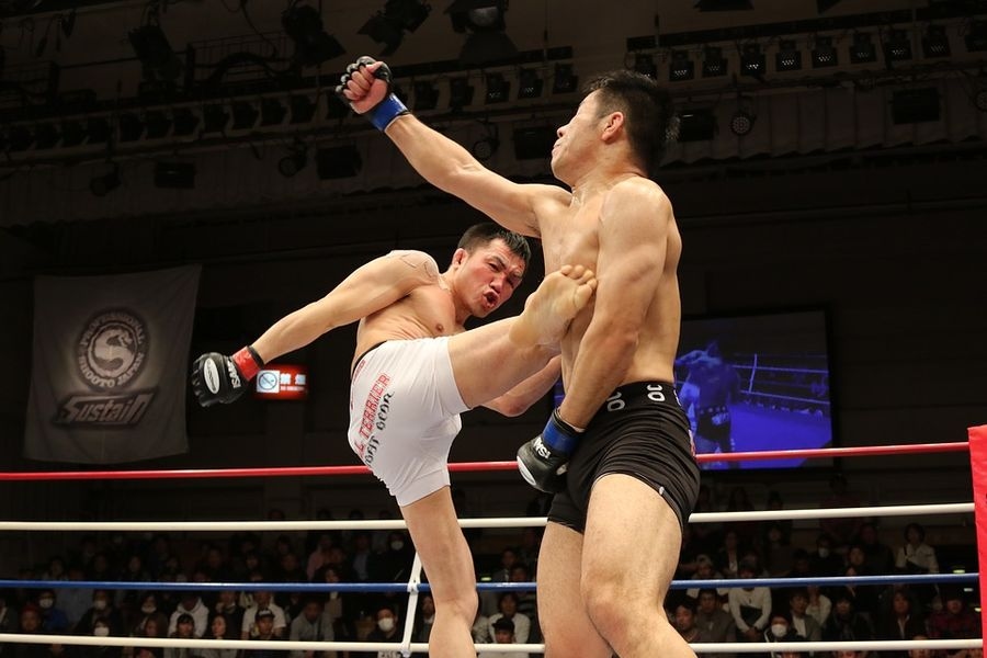 Combattant en short blanc portant un coup de pied circulaire au corps de son adversaire sur un ring de MMA au Japon, en gants ouverts